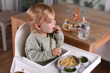 Yüksek sandalyede oturan bir çocuk spagetti ve bezelye yiyor. Bebek menüsü, çocuk menüsü, eğlenceli keşifler, günlük çocukluk rutinleri, kendini besleme, bebek sütten kesilmeye yol açtı..
