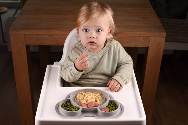 A child sitting in a high chair eating spaghetti and peas. Baby menu, toddler's mealtime alongside playful exploration, everyday childhood routines, self feeding, BLW baby led weaning.