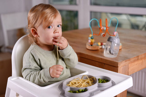 A child sitting in a high chair eating spaghetti and peas. Baby menu, toddler's mealtime alongside playful exploration, everyday childhood routines, self feeding, BLW baby led weaning.