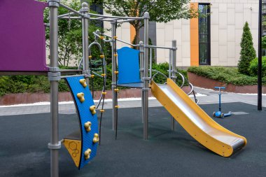 Çağdaş binalar ve yeşilliklerin bulunduğu bir yerleşim bölgesinde, tırmanma yapıları ve slaytların yer aldığı açık hava oyun alanı