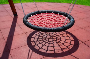 Kırmızı bir ağ salıncağı, açık hava parkında canlı renkler ve geometrik şekillerle görsel ilgi oluşturan oyun alanı fayansları üzerinde karmaşık bir gölge oluşturur.