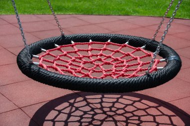 Kırmızı bir ağ salıncağı, açık hava parkında canlı renkler ve geometrik şekillerle görsel ilgi oluşturan oyun alanı fayansları üzerinde karmaşık bir gölge oluşturur.