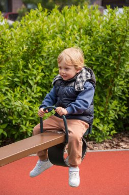 Güneşli bir parkta tahterevalli sürüşünün keyfini çıkaran bir çocuk. Ortam renkli bir oyun alanını çevreleyen ve boş zamanlarında neşe ve bağlılık tasvir eden doğayı içeriyor.
