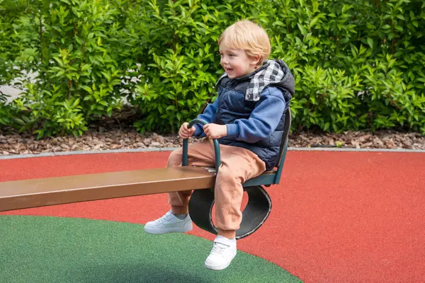 Güneşli bir parkta tahterevalli sürüşünün keyfini çıkaran bir çocuk. Ortam renkli bir oyun alanını çevreleyen ve boş zamanlarında neşe ve bağlılık tasvir eden doğayı içeriyor.