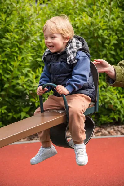 Güneşli bir parkta tahterevalli sürüşünün keyfini çıkaran bir çocuk. Ortam renkli bir oyun alanını çevreleyen ve boş zamanlarında neşe ve bağlılık tasvir eden doğayı içeriyor.
