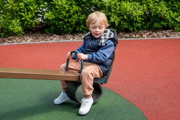 Güneşli bir parkta tahterevalli sürüşünün keyfini çıkaran bir çocuk. Ortam renkli bir oyun alanını çevreleyen ve boş zamanlarında neşe ve bağlılık tasvir eden doğayı içeriyor.