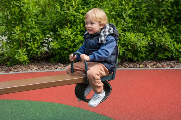 Güneşli bir parkta tahterevalli sürüşünün keyfini çıkaran bir çocuk. Ortam renkli bir oyun alanını çevreleyen ve boş zamanlarında neşe ve bağlılık tasvir eden doğayı içeriyor.