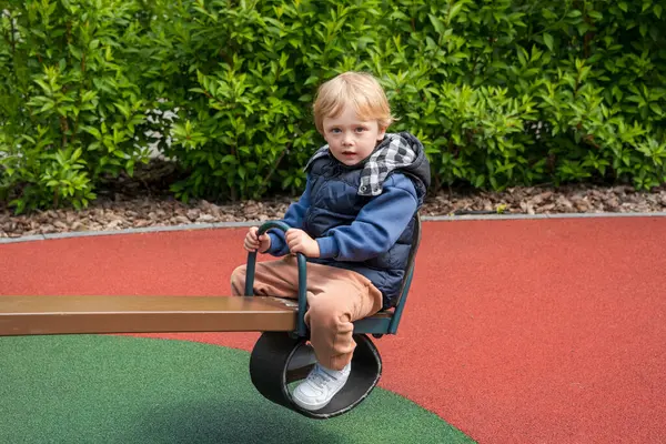 Güneşli bir parkta tahterevalli sürüşünün keyfini çıkaran bir çocuk. Ortam renkli bir oyun alanını çevreleyen ve boş zamanlarında neşe ve bağlılık tasvir eden doğayı içeriyor.