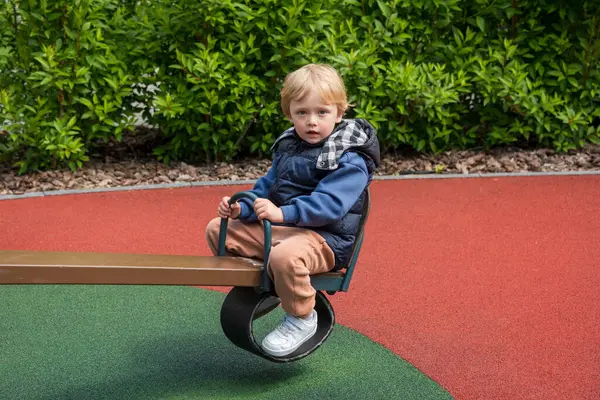 Güneşli bir parkta tahterevalli sürüşünün keyfini çıkaran bir çocuk. Ortam renkli bir oyun alanını çevreleyen ve boş zamanlarında neşe ve bağlılık tasvir eden doğayı içeriyor.