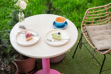 İki renkli tatlı, bir fincan kahve ve bir vazo çiçeğin bulunduğu rahat bir açık kahve masası, canlı bir bahçe arka planına kurulmuş, keyifli ve rahatlatıcı bir atmosfer sunuyor..