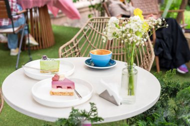 İki renkli tatlı, bir fincan kahve ve bir vazo çiçeğin bulunduğu rahat bir açık kahve masası, canlı bir bahçe arka planına kurulmuş, keyifli ve rahatlatıcı bir atmosfer sunuyor..