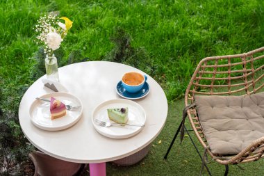 İki renkli tatlı, bir fincan kahve ve bir vazo çiçeğin bulunduğu rahat bir açık kahve masası, canlı bir bahçe arka planına kurulmuş, keyifli ve rahatlatıcı bir atmosfer sunuyor..