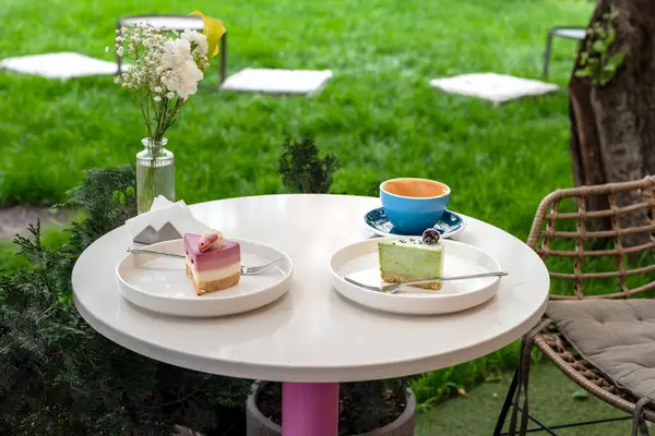 İki renkli tatlı, bir fincan kahve ve bir vazo çiçeğin bulunduğu rahat bir açık kahve masası, canlı bir bahçe arka planına kurulmuş, keyifli ve rahatlatıcı bir atmosfer sunuyor..