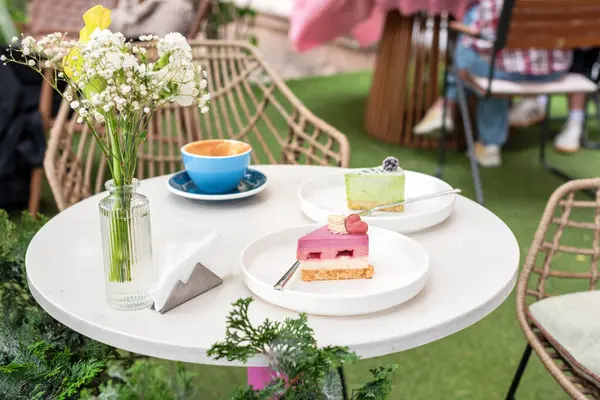 İki renkli tatlı, bir fincan kahve ve bir vazo çiçeğin bulunduğu rahat bir açık kahve masası, canlı bir bahçe arka planına kurulmuş, keyifli ve rahatlatıcı bir atmosfer sunuyor..