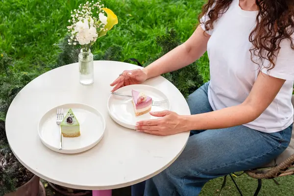 Açık havada oturan bir kadın, yeşil bir vazoyla çevrili beyaz bir masada canlı tatlılar yemekten hoşlanır. Görüntü huzurlu, rahatlatıcı ve hoşgörülü bir atmosfer yayar.