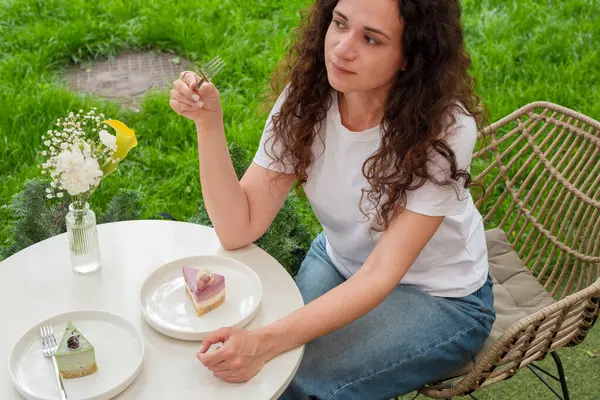 İki tabak tatlısıyla açık bir masada oturan bir kadın, huzurlu yeşil bir bahçede manzaranın ve sakin çevrenin tadını çıkarıyor. Düşünceli bir şekilde gülümsüyor, doğadaki rahatlık ve neşeyi simgeliyor.