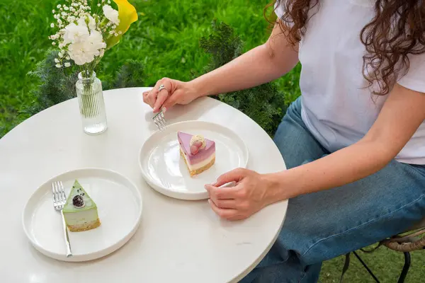 Açık havada oturan bir kadın, yeşil bir vazoyla çevrili beyaz bir masada canlı tatlılar yemekten hoşlanır. Görüntü huzurlu, rahatlatıcı ve hoşgörülü bir atmosfer yayar.