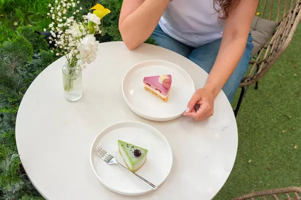 Açık havada oturan bir kadın, yeşil bir vazoyla çevrili beyaz bir masada canlı tatlılar yemekten hoşlanır. Görüntü huzurlu, rahatlatıcı ve hoşgörülü bir atmosfer yayar.