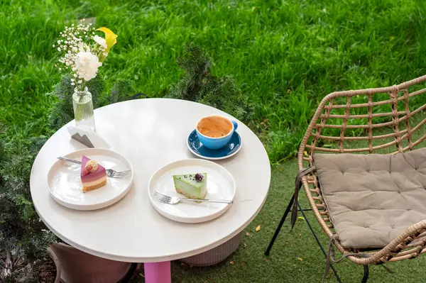 İki renkli tatlı, bir fincan kahve ve bir vazo çiçeğin bulunduğu rahat bir açık kahve masası, canlı bir bahçe arka planına kurulmuş, keyifli ve rahatlatıcı bir atmosfer sunuyor..
