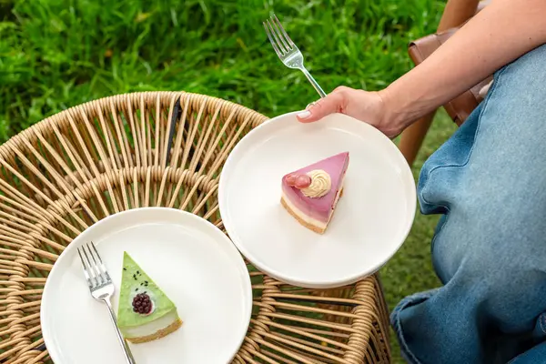 Bir kadın bahçe düzeninde otururken renkli katmanlı bir pasta tabağı tutuyor. Yumuşak ışıklandırma ve yemyeşil arka plan sahnenin taze ve neşeli havasını arttırır.