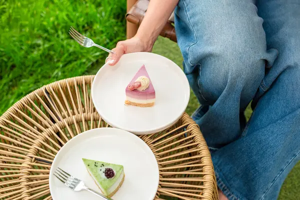 Bir kadın bahçe düzeninde otururken renkli katmanlı bir pasta tabağı tutuyor. Yumuşak ışıklandırma ve yemyeşil arka plan sahnenin taze ve neşeli havasını arttırır.