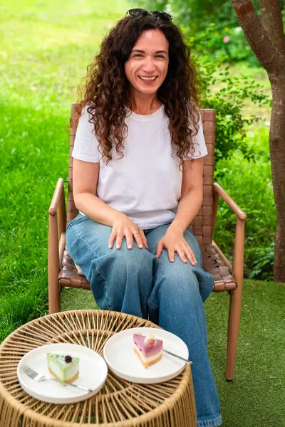 İki tabak tatlısıyla açık bir masada oturan bir kadın, huzurlu yeşil bir bahçede manzaranın ve sakin çevrenin tadını çıkarıyor. Düşünceli bir şekilde gülümsüyor, doğadaki rahatlık ve neşeyi simgeliyor.