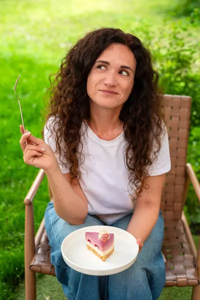 İki tabak tatlısıyla açık bir masada oturan bir kadın, huzurlu yeşil bir bahçede manzaranın ve sakin çevrenin tadını çıkarıyor. Düşünceli bir şekilde gülümsüyor, doğadaki rahatlık ve neşeyi simgeliyor.