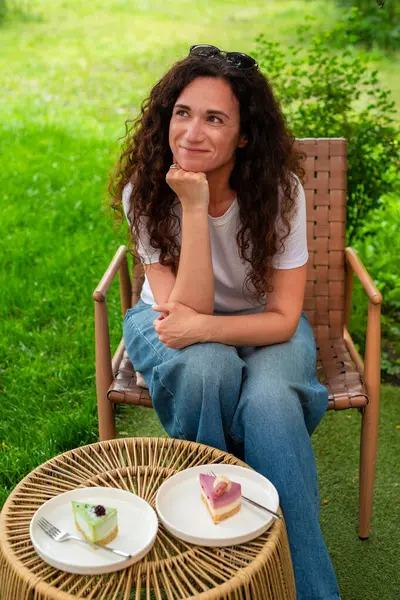 İki tabak tatlısıyla açık bir masada oturan bir kadın, huzurlu yeşil bir bahçede manzaranın ve sakin çevrenin tadını çıkarıyor. Düşünceli bir şekilde gülümsüyor, doğadaki rahatlık ve neşeyi simgeliyor.