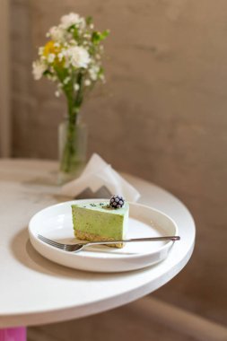 Böğürtlen soslu bir dilim yeşil tatlı, beyaz bir tabakta zarif bir şekilde sunuluyor, yanında en az arka plan ve çiçekli süsleme, modern ve estetik gıda tasarımı sunuyor.