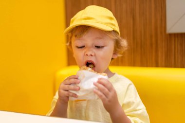 Sarı şapka ve gömlek giyen genç bir çocuk canlı bir restoranda çizburger yiyor, sıradan bir yemek ortamında fast food yemekten zevk alıyor. Çocuk diyeti, kötü yemek seçenekleri, erken dönem abur cubur bağımlılığı..