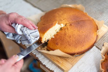 Close-up view of a person slicing a tender, golden-brown cake, freshly baked and placed on parchment paper over a wooden board, creating a cozy and warm kitchen atmosphere