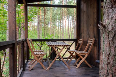 Sakin bir orman manzarasına bakan bir balkona yerleştirilmiş, ahşap bir masa ve sandalyeler. Sahne dinginliği, doğanın güzelliğini ve rahatlamak için ideal dış mekanların çekiciliğini yansıtıyor.