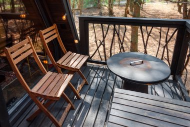 Dışarıdaki güvertede iki tahta sandalye ve bir yuvarlak masa var, doğal orman alanıyla çevrili. Doğanın tadını çıkarmak, rahatlamak, açık hava yemekleri, kulübe inzivaları, orman kaçamakları, ya da küçük evler.