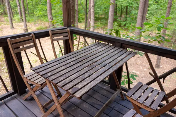 Sakin bir orman manzarasına bakan bir balkona yerleştirilmiş, ahşap bir masa ve sandalyeler. Sahne dinginliği, doğanın güzelliğini ve rahatlamak için ideal dış mekanların çekiciliğini yansıtıyor.