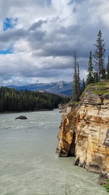 View over Athabasca River flowing from nearby Athabasca Falls, Jasper National Park, AB, Canada, with white-water raft in distance on river and river shore with colorful rock formations of limestone clipart