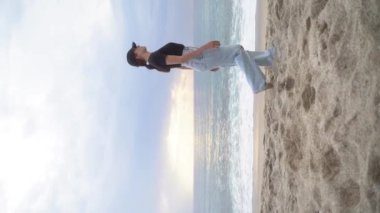 Dikey video. Makaralar. Genç bir kadın kıyı şeridi boyunca yürüyor ve arkaplanı deniz olan çerçeveden çıkıyor. Serbest Yaşam Tarzı Yavaş Hareket