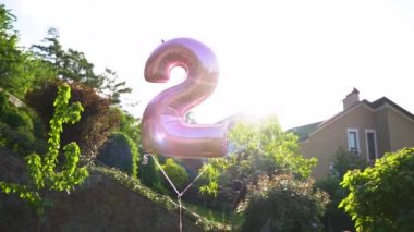 Yeşil ağaçların arka planında uçan iki numaralı pembe bir balon. Tatiller, yıldönümleri, doğum günleri için parlak bir ekran koruyucu. Yavaş Hareket 120