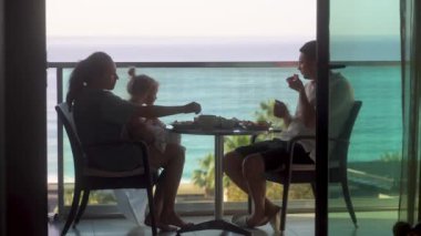 Üç kişilik genç bir aile açık hava balkonunda inanılmaz deniz manzaralı kahvaltı yapıyorlar. Mükemmel bir sabah. Otelde. Alanya Türkiye 2021 Yüksek kaliteli 4K görüntü