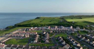 Okyanus kıyısındaki güzel özel evlerin üzerinde bir deniz feneri zeminine karşı uçmak. Yeşil vadi. Yaz. Gün batımı. Waterford, Tramore, İrlanda. Yüksek kalite 4k görüntü 