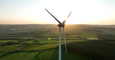 Havadan. Dairesel. Gün batımına karşı bıçakları olan büyük bir yel değirmeni. Yeşil İrlanda ormanları. Alternatif enerji. Temiz enerji. Yüksek kalite 4k