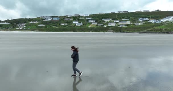 一个女人走在荒芜的海滩上 孤独和寒冷 一个荒芜的海滩镜像喷出脚下 一个关于海洋的观点 桑迪海滩蓝水 多云的天气 野生大西洋方式 — 图库视频影像