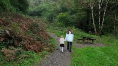 Bir anne ve çocuğu park alanında yürürler. Anne, çocuğun elini tutuyor. Bir anne ve çocuğu bir tepe boyunca yürüyorlar. Turistik bir yer. Ormanda bir yürüyüş. Yürüyüş zamanı.