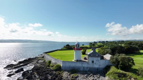緑の海岸の背景にある白い灯台の上を飛行してください 白い灯台が付いている絵の海岸線 アイルランドのウォーターフォード郡にあるバリナックアワーティ灯台の空中眺望 — ストック動画