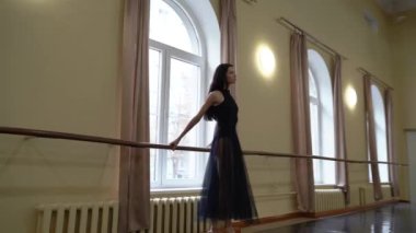 Profesyonel bir balerin, bale salonunun penceresinin önündeki bale makinesinde dans eder. Güzel dans figürleri gösteriyor. Zarif balerin, büyük bir salonda klasik balerin unsurlarıyla dans eder.