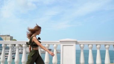 Yazlık takım elbiseli genç bir kadın arka planda deniz varken koşup zıplıyor. Denize hayranım. Moda renkli etek. Denize karşı güzel bir kız. Yaz tatili. Şehir ve deniz manzarası