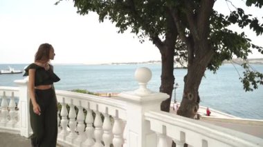 Yazlık takım elbiseli genç bir kadın arka planda denizle birlikte arka planda geziniyor. Ağır çekim. Moda renkli etek, büyük şapka. Denize karşı güzel bir kız. Kız.