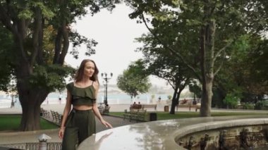 Yazlık takım elbiseli bir kız yazın çeşmenin yanında yürür. Moda renkli etek, büyük şapka. Denize karşı güzel bir kız. Kız saçını düzeltiyor. Yaz tatili. Parkta yaz. Kadın