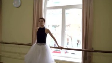 Profesyonel bir balerin, büyük bir bale salonunun penceresinin önünde bir bale barında dans eder. Güzel dans figürleri gösteriyor. Zarif balerin büyük bir salonda klasik balerin unsurlarıyla dans eder. Dans et
