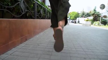 Kapatın. Yeşil bir şehirde caddede koşan bir kadın. Açık yeşil renk pantolon geliştirme. Kızların bacaklarını kapat. Şehirde sandaletleriyle koşan bir kız. Yaz havası. Yavaş Hareket