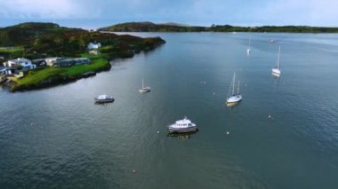 Bereketli yeşil tepelerle çevrili sakin sularda süzülen yelkenli teknelerinin olduğu sakin bir liman sahnesi. İrlanda 'da küçük bir kasabanın arka planında beyaz yatlar ve tekneler var. Kıyıda bir balıkçı kasabası.